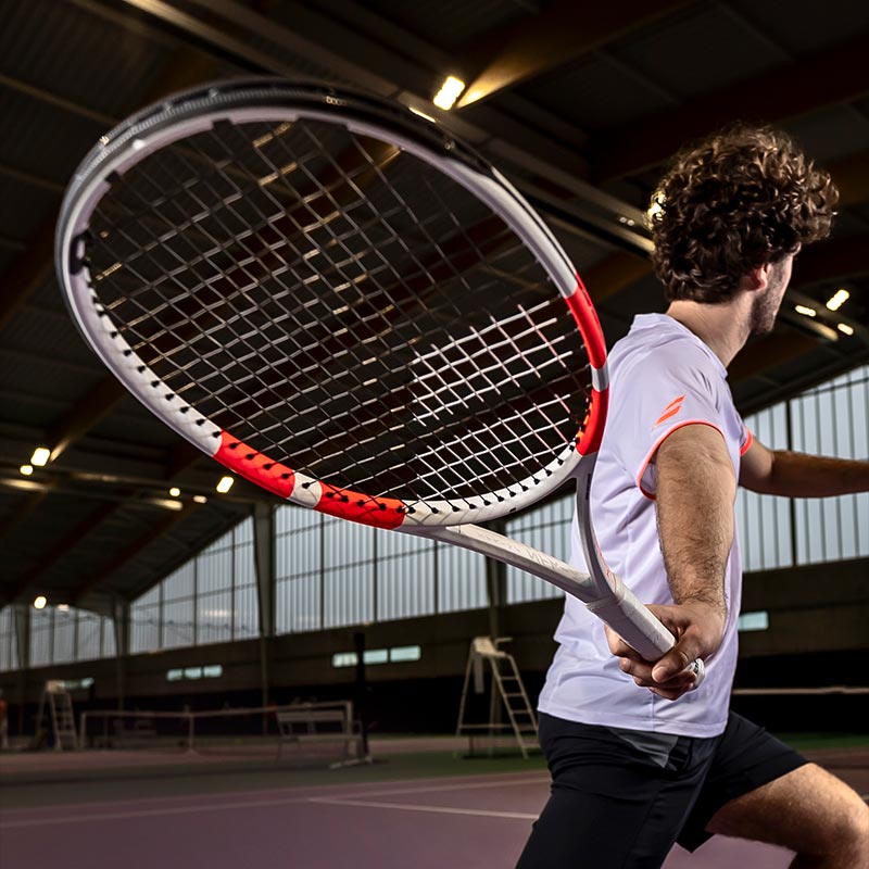 Lifestyle image: Mid-swing shot of a man playing with a Babolat Pure Strike 2024 tennis racquet on a dimly lit indoor court.